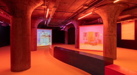 Intérieur de style industriel avec des colonnes en béton, des tuyaux de plafond apparents, un éclairage chaud orange-rouge et des projections multimédias présentant des installations artistiques dans un espace d’exposition moderne.  Crédit image: Alex Da Corte, le musée Mouse (oreille de Van Gogh), 2024. Ver d’oreille, vue d’installation, MOCA Toronto, 2024. Photographe: LF Documentation.