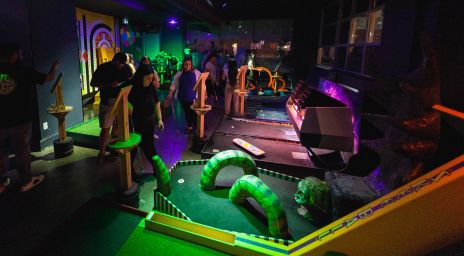 Terrain de mini-golf intérieur avec des obstacles colorés en forme de serpent, un éclairage à intensité réduite et des gens jouant ou regardant dans une atmosphère vibrante et ludique.