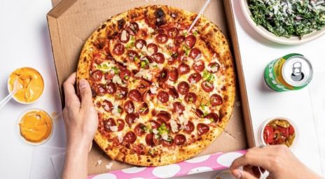 Une personne tenant une part de pizza au pepperoni avec des poivrons verts et du fromage provenant d’une boîte à pizza, accompagnée de sauces à tremper, d’un bol de salade, d’une canette de soda et d’un contenant de légumes marinés rouges.