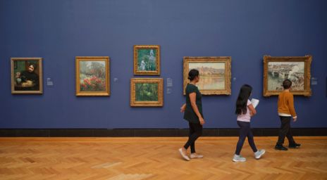 Galerie d’art avec des tableaux encadrés sur un mur bleu et des visiteurs qui passent.