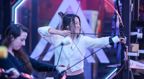 Deux personnes pratiquant le tir à l’arc à l’intérieur avec un éclairage moderne.