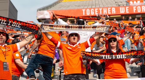 Forge FC fans in orange shirts cheering in stadium stands, holding black-and-orange scarves that read “’Til I Die” and “We Make This Look Easy.”