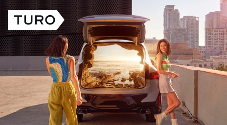 Two women and a hatchback with an oceanside sunset visible through the tailgate